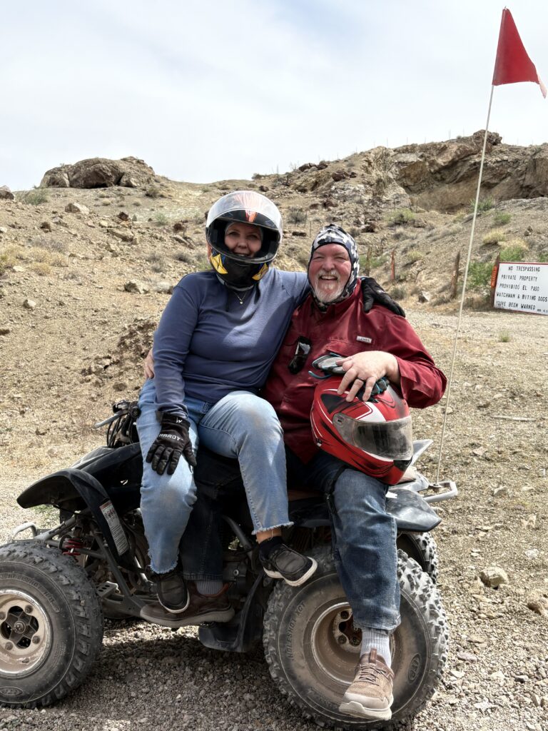 Two people enjoy an ATV tour near Las Vegas, Nevada, smiling in helmets with a red flag and warning sign in the desert landscape.