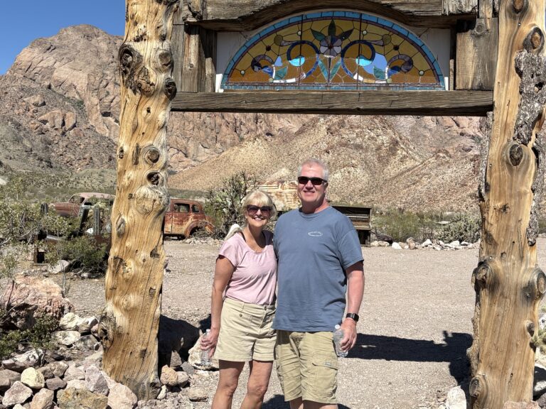 Smiling couple under rustic arch with stained glass in Nevada ghost town, desert hills, and old vehicles near Las Vegas.