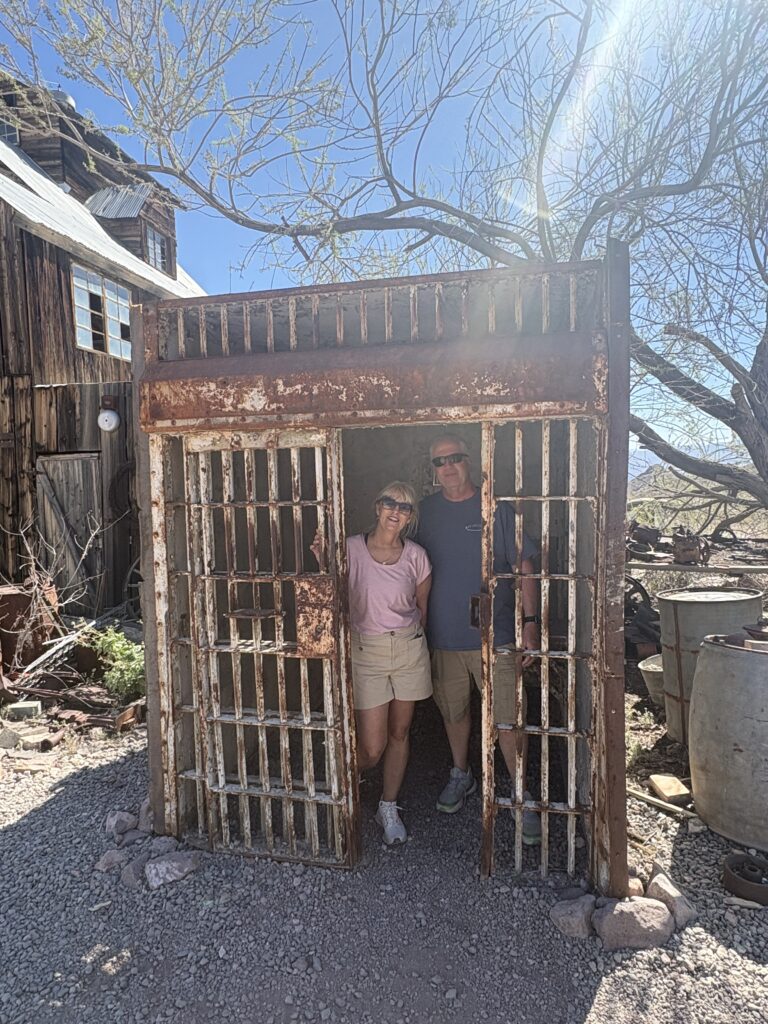 Smiling visitors in a rusty outdoor jail cell at a Nevada Ghost Town near Las Vegas, surrounded by desert and vintage buildings.