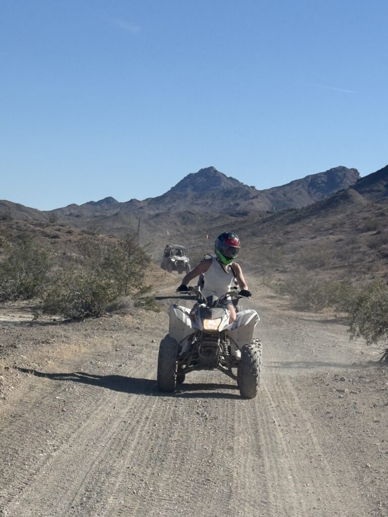 ATV rider in helmet explores a rocky Nevada desert trail near Las Vegas, with mountains and an ATV tour group following behind.