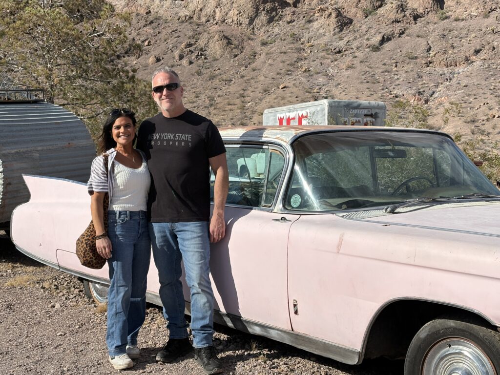 Smiling couple by vintage pink car near rocky Nevada hills and a silver trailer, perfect for Las Vegas Ghost Town sightseeing.
