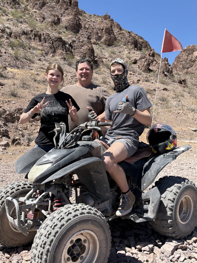Group posing by a black ATV in Nevada desert near Las Vegas, ready for exciting ATV tour with a red flag on rocky terrain.