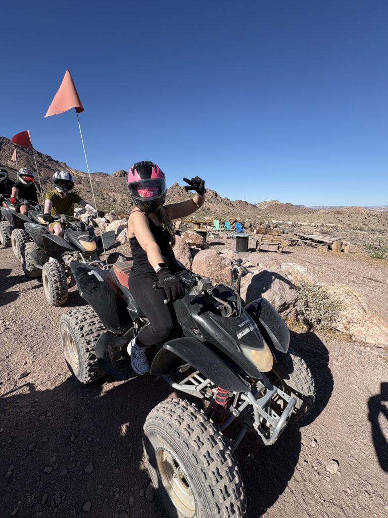 Rider in pink helmet gives thumbs-up on ATV tour near Las Vegas, Nevada; others follow on desert trail under blue sky.