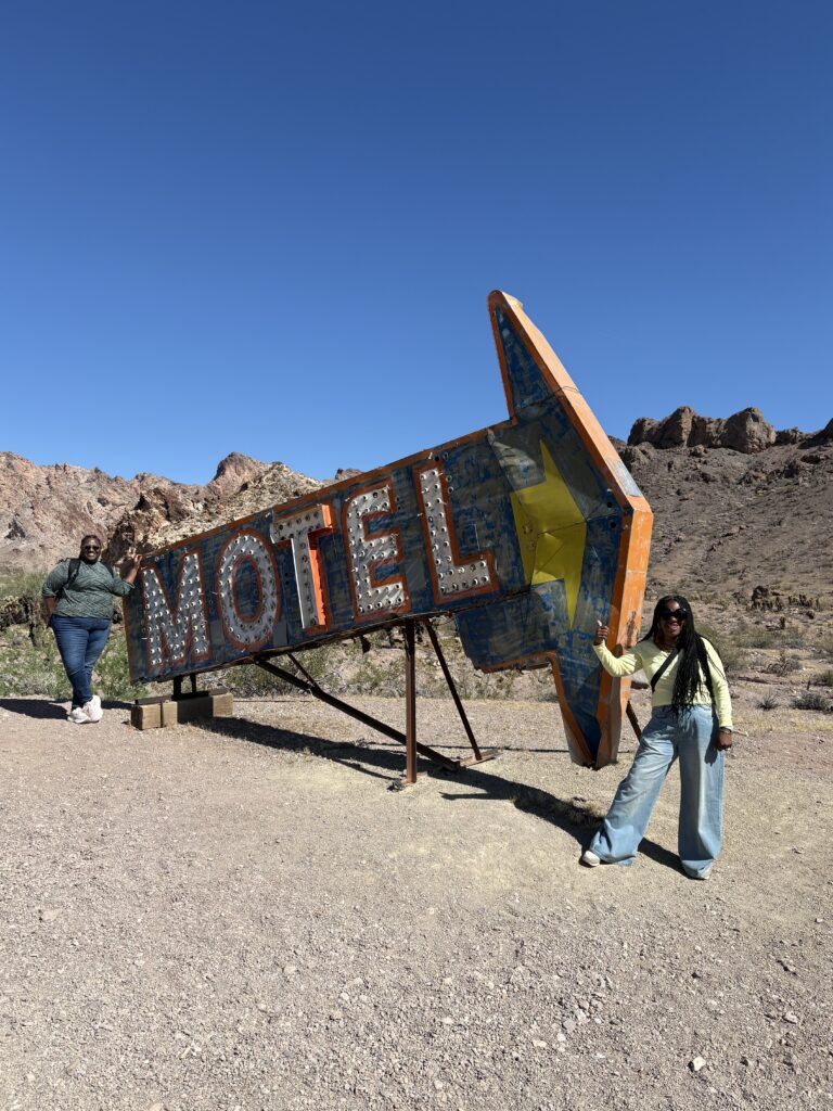 Two people by a vintage arrow-shaped MOTEL sign in Nevada desert near Las Vegas, rocky hills and blue sky. Ghost Town sightseeing nearby.