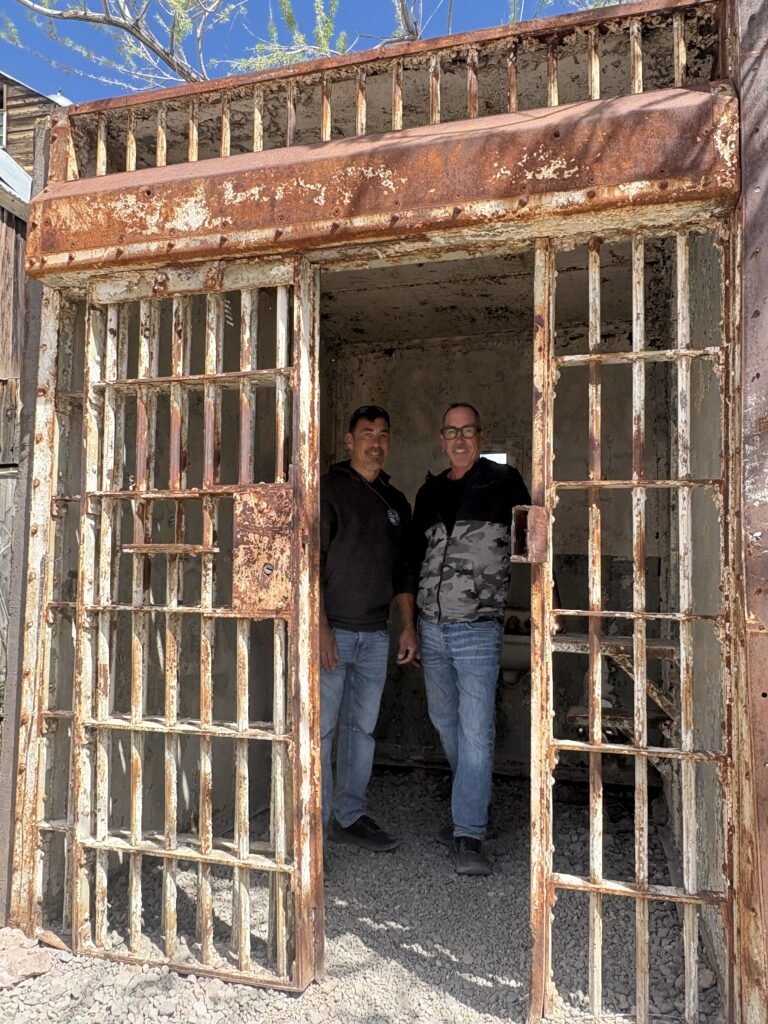 Two men smile inside an old rusty Nevada jail cell, sunlight shining in—perfect for Las Vegas ghost town sightseeing tours.