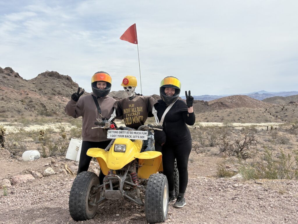 ATV tour near Las Vegas: two riders and a skeleton on a yellow quad in Nevada desert, rocky hills, ghost town sightseeing vibes.