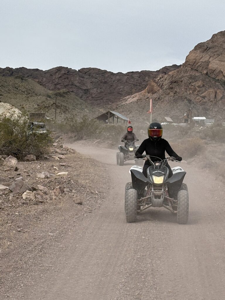 Two riders in helmets enjoy an ATV tour on a dusty Nevada desert trail near Las Vegas with mountains in the background.
