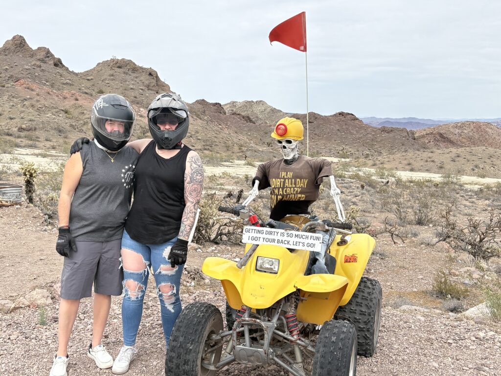 Adventurers in helmets pose by a yellow ATV with skeleton driver near Las Vegas, Nevada desert, perfect for ATV tours.