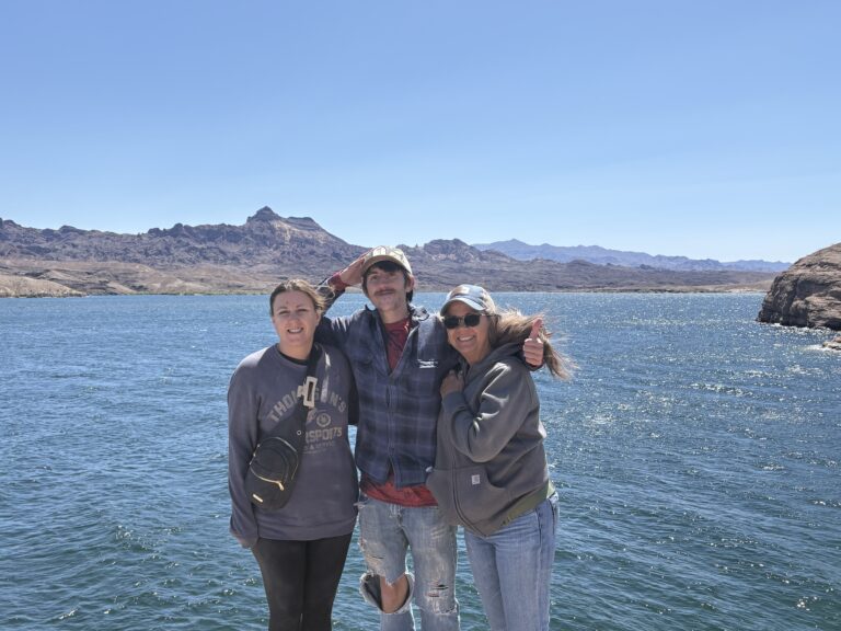 Three friends smile by the Colorado River near Nevada mountains, dressed for ATV tours under clear skies near Las Vegas.