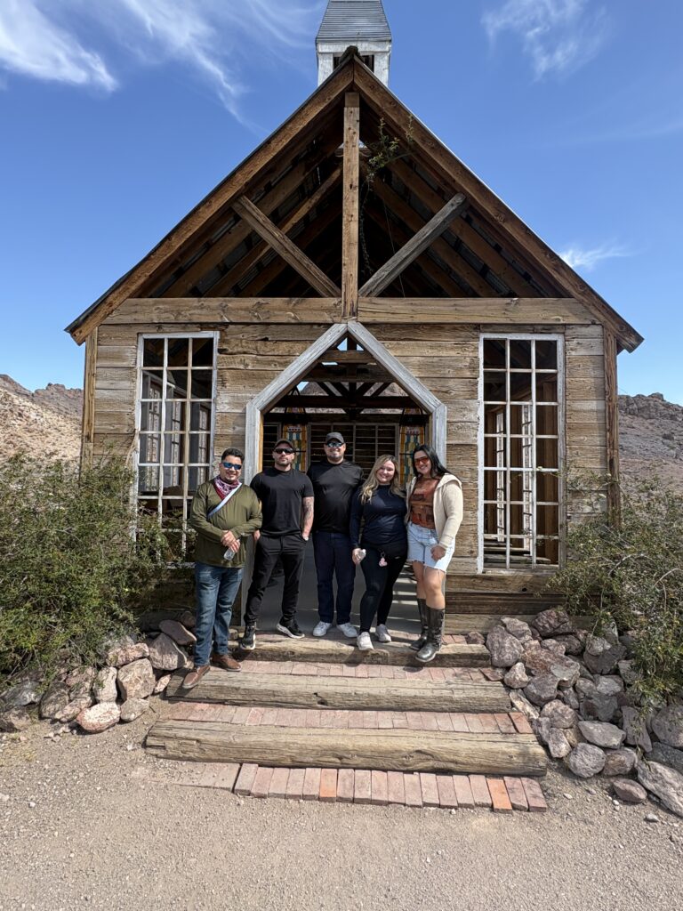 Five visitors smile in front of a rustic Nevada ghost town near Las Vegas, surrounded by desert plants and rocky terrain.