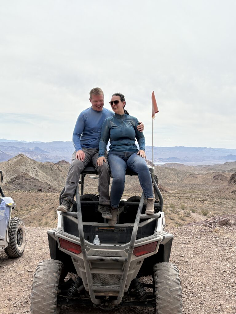 A couple smiles on a RZR during an ATV tour near Las Vegas, Nevada, with desert mountains and a flag in the scenic background.
