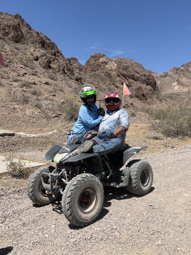 Two riders in helmets on an ATV tour explore a rocky Nevada desert near Las Vegas with red flags and mountains behind.