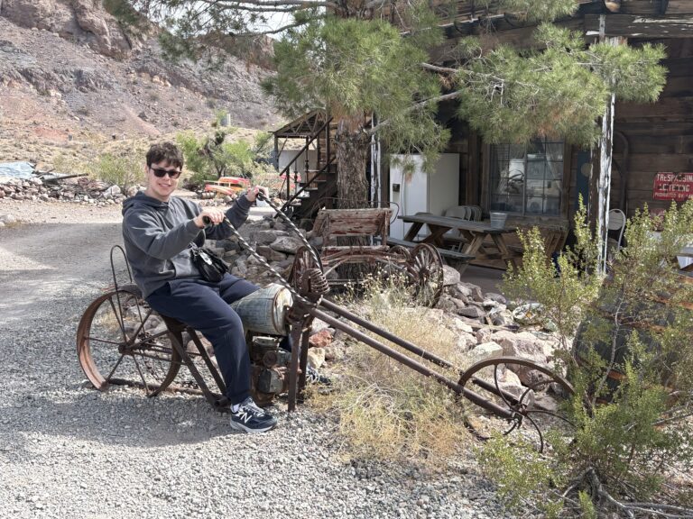 Smiling visitor on a vintage motorcycle sculpture outside a rustic building in Nevada Ghost Town near the Colorado River.