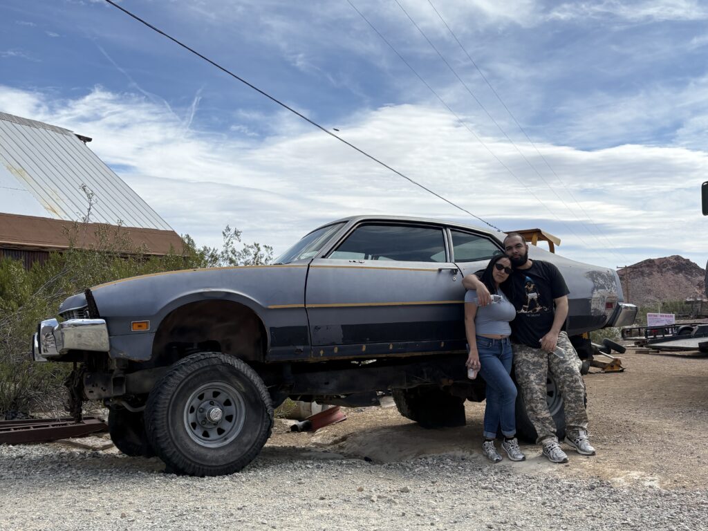 Smiling couple poses by a rugged off-road car in Nevada desert, near Las Vegas, with ghost town and mountains in background.