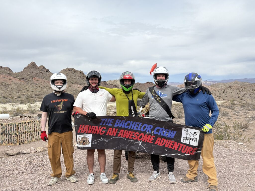 Group in helmets and gear poses with "The Bachelor Crew" banner during Nevada ATV tour near Las Vegas desert and Colorado River.