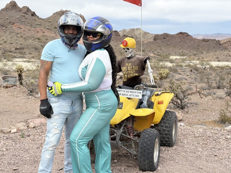 Couple in riding gear poses by yellow ATV with skeleton in helmet near Nevada desert hills, perfect for Las Vegas ATV tours.