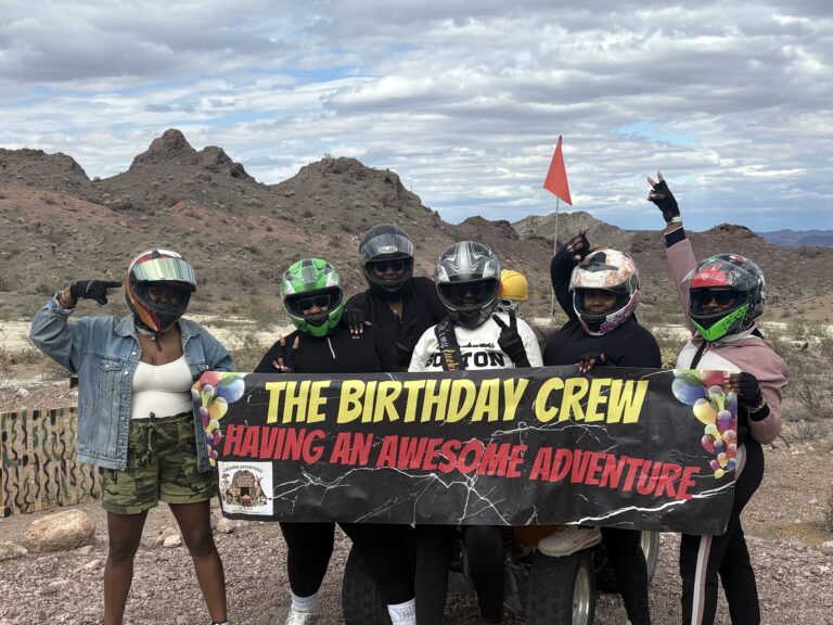 Six friends in helmets pose by Nevada desert hills with a birthday banner, ready for an epic Las Vegas ATV tour adventure.
