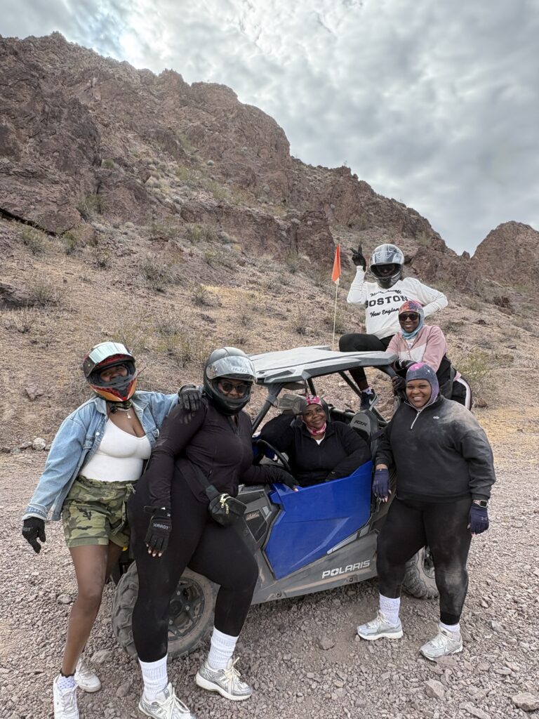 Five women in helmets pose by a Polaris RZR during an ATV tour near Las Vegas’s rocky cliffs and cloudy Nevada sky.