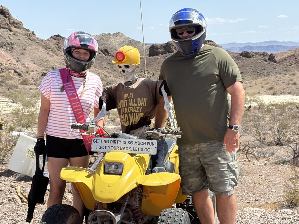 Three helmeted riders and a skeleton pose by a yellow ATV in the Nevada desert near Las Vegas ghost town ATV tours.