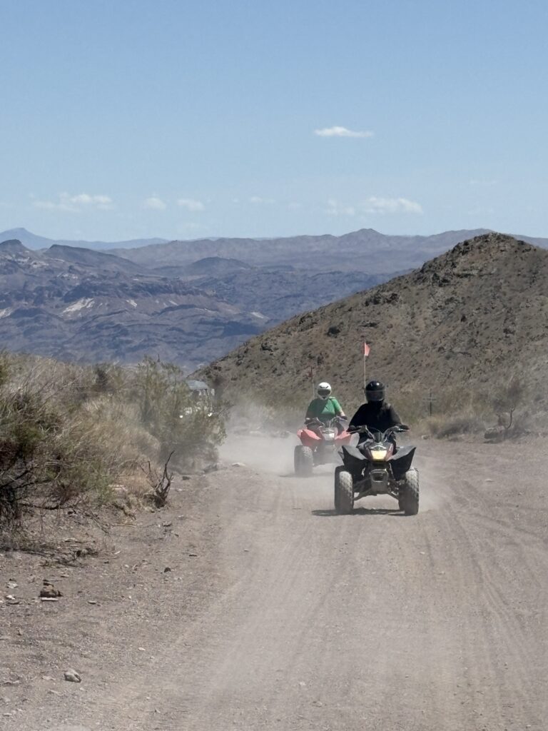 Adventurers on an ATV tour ride along a dusty Nevada desert trail near the Colorado River, mountains, and blue sky in view.