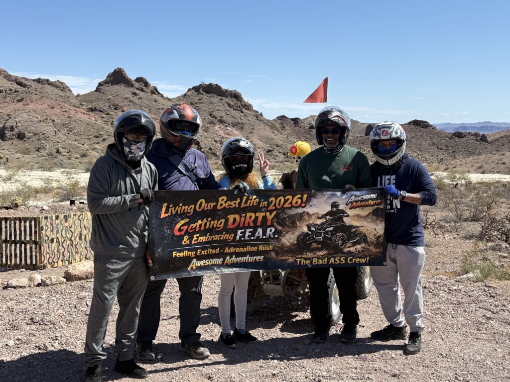 Group in helmets on rocky Nevada terrain, holding adventure banner near ATV with Las Vegas, Colorado River & mountains behind.
