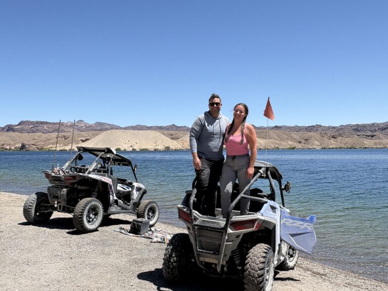 Two people enjoy an ATV tour by the Colorado River near Las Vegas, Nevada, with RZR vehicles and scenic rocky hills in the background.