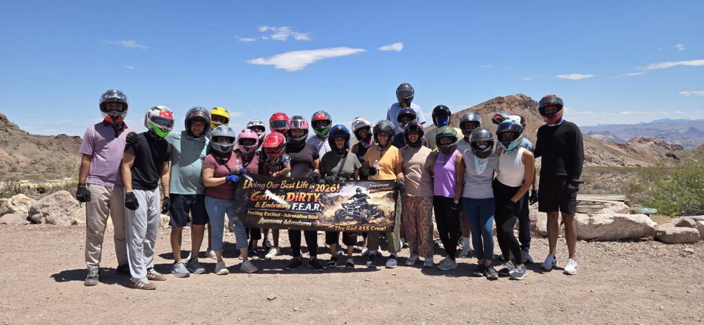Adventurers in helmets hold a "Live your best life" banner on rocky Nevada terrain near Las Vegas, ready for an ATV or RZR tour.