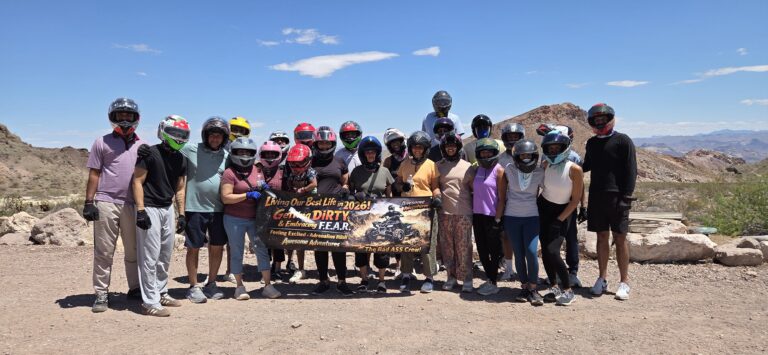Adventurers in helmets hold a "Live your best life" banner on rocky Nevada terrain near Las Vegas, ready for an ATV or RZR tour.