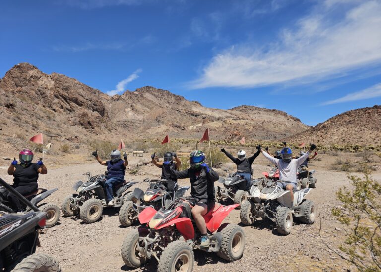Helmeted riders on ATVs raise arms, posing in Nevada desert with red flags—Las Vegas ATV tour near Colorado River mountains.
