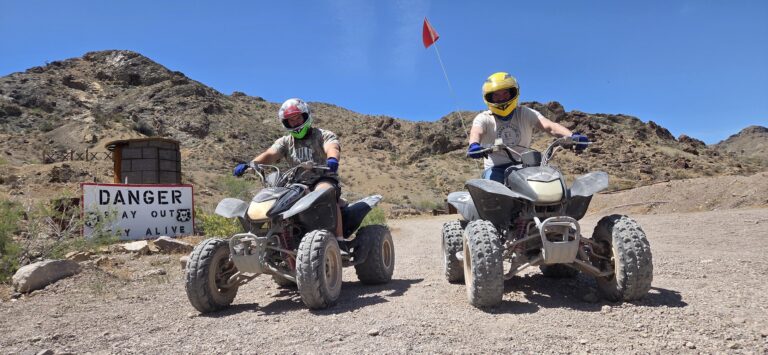 Adventurers ride ATVs on a Nevada desert trail near Las Vegas; “DANGER STAY OUT!” sign warns by the clear blue sky.