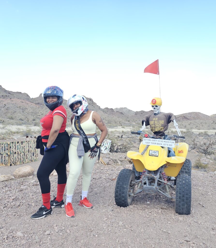 Two women pose by a yellow ATV in the Nevada desert near Las Vegas, ready for an exciting ATV tour with rocky hills behind them.