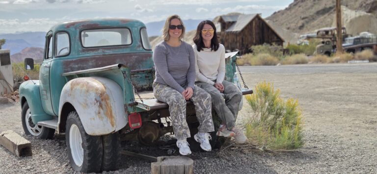 Two women smile on a vintage turquoise pickup in a Nevada ghost town near Las Vegas, perfect for ATV tours and sightseeing.
