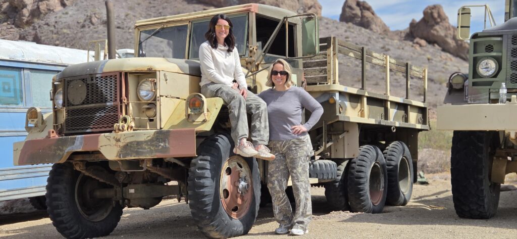 Two women in camo by a vintage military truck smile near Nevada’s rocky desert, perfect for Las Vegas ATV tours or Ghost Town trips.