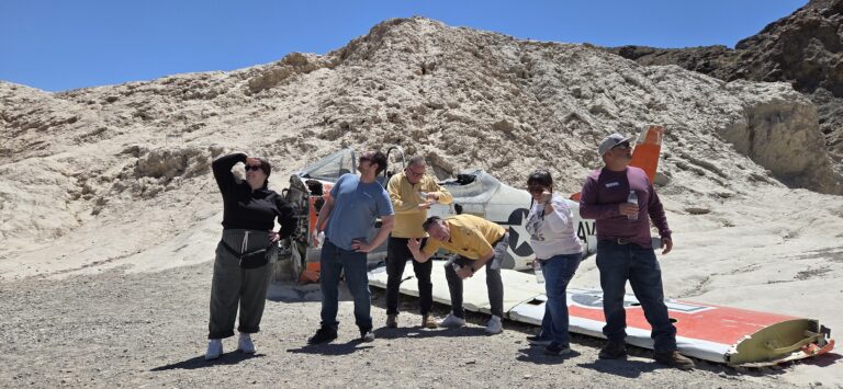 Group posing by a crashed airplane in Nevada’s rocky desert near Las Vegas, with playful energy and scenic hills behind.
