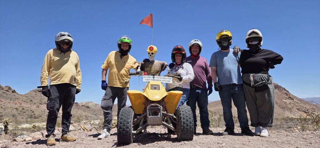 Group in helmets poses by yellow ATV during Las Vegas Nevada desert ATV tour, rocky mountains and blue sky in the background.