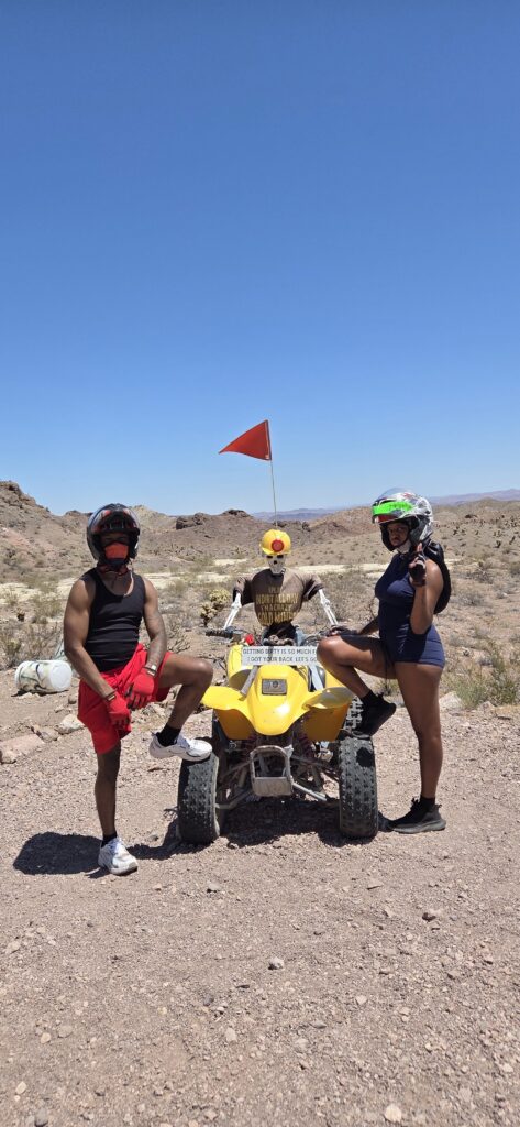 Three helmeted adventurers with a yellow ATV in the Nevada desert, ready for Las Vegas ATV tours. Red flag, rocky hills behind.