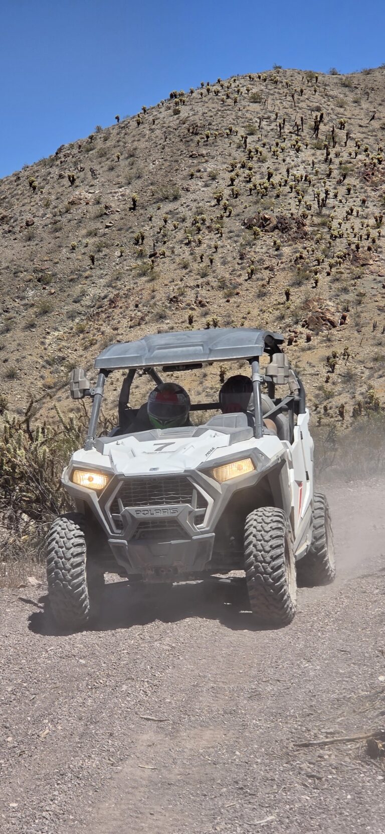 A white RZR off-road vehicle with two helmeted riders explores a dusty Nevada desert trail near Las Vegas and the Colorado River.