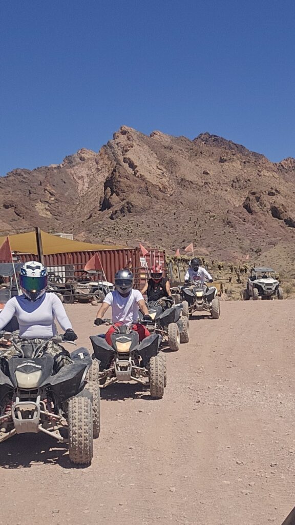 ATV tour near Las Vegas: four riders with helmets cruise a desert trail by rocky Nevada mountains, tents, and vehicles nearby.