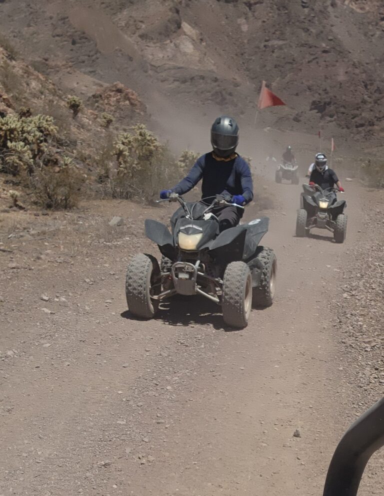 Riders on an ATV tour near Las Vegas traverse a dusty desert trail, red flags flying, with Nevada hills in the background.