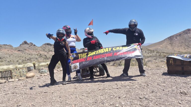 Five riders in helmets pose in a Nevada desert near Las Vegas, holding "The Birthday Crew" banner during an ATV tour under blue skies.
