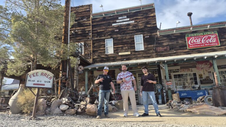 Three men point at a Fee Area sign at a Nevada ghost town near Las Vegas, surrounded by antiques and desert plants.