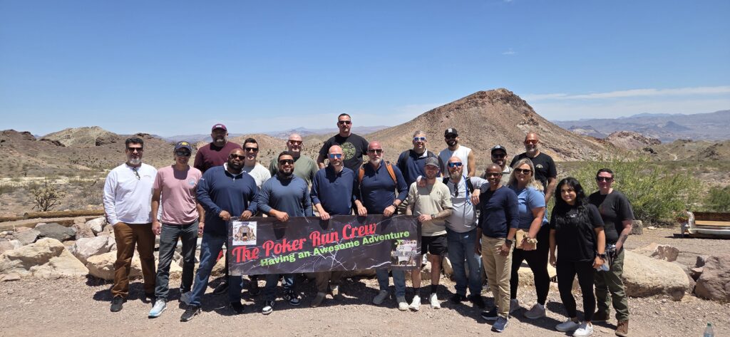 Group of 20 poses by Nevada rocky hills with "Poker Run Crew" banner, enjoying ATV tours near Las Vegas and Colorado River.