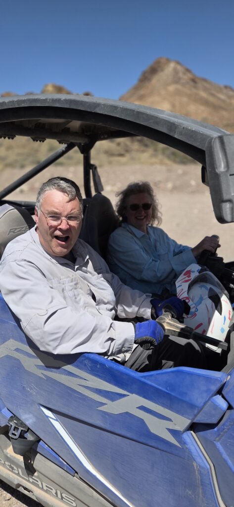 Smiling adults in a blue RZR on a Nevada desert ATV tour near Las Vegas, with rocky hills and clear blue sky in the background.