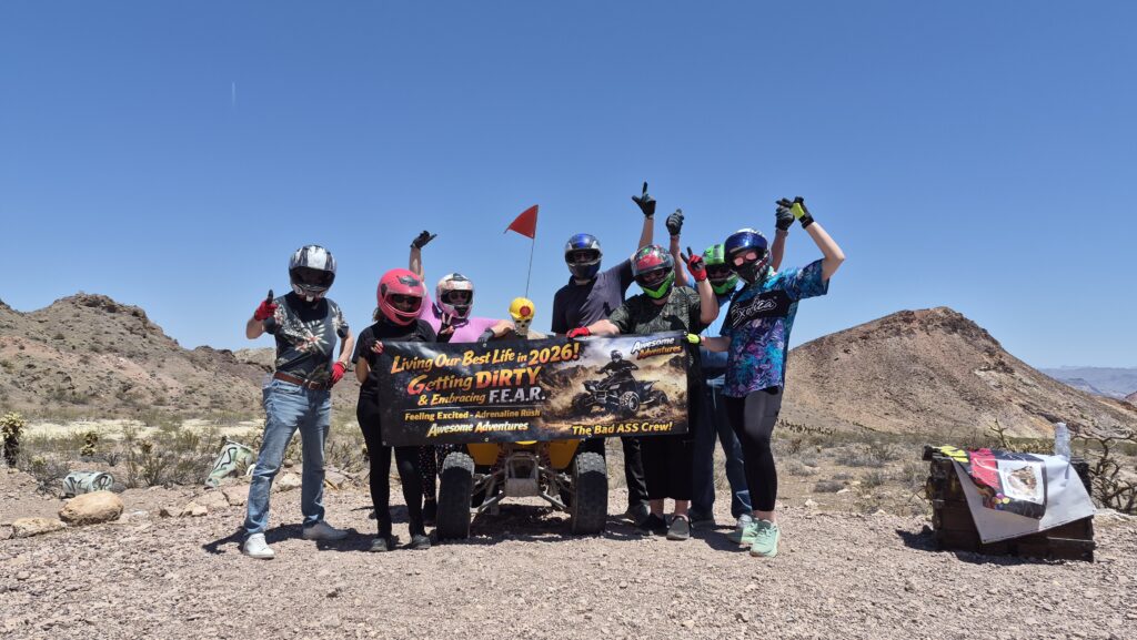 Six people in helmets pose by an ATV and banner in Nevada’s desert, ready for Las Vegas RZR off-road and ghost town tours.