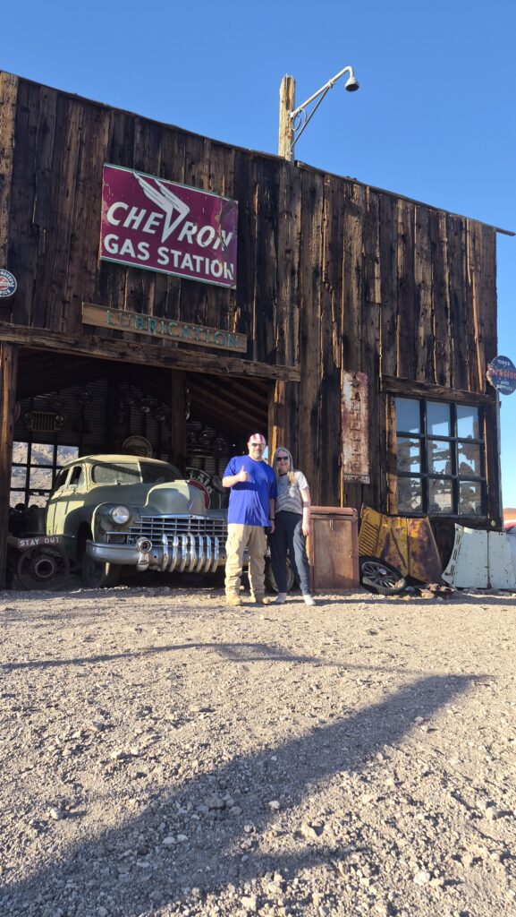 Couple at Nevada ghost town near Las Vegas, with vintage Chevron sign, classic car, and gas pumps—perfect for sightseeing tours.