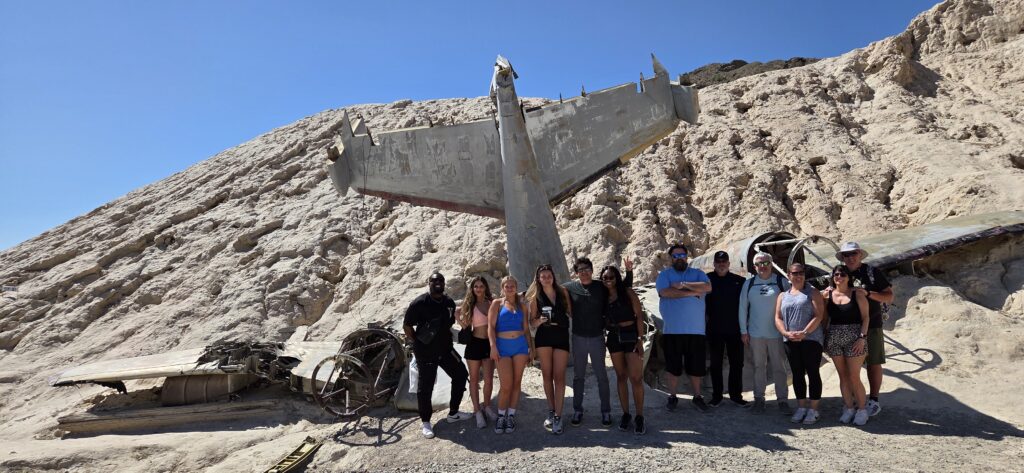 Visitors pose by rocky Nevada hills and aircraft wreckage during an ATV tour near Las Vegas under a clear blue sky.