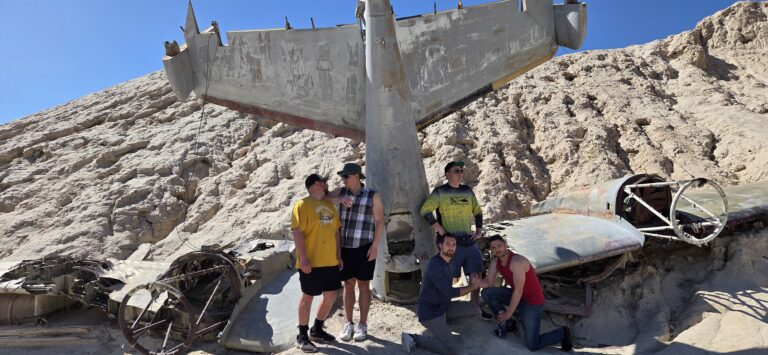 Six adventurers pose by a weathered plane wreck on a Nevada ATV tour near Las Vegas, under clear blue skies.