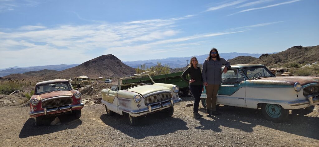 Two people stand among vintage cars in a sunny Nevada desert near Las Vegas, with mountains and blue sky—perfect for Ghost Town tours.