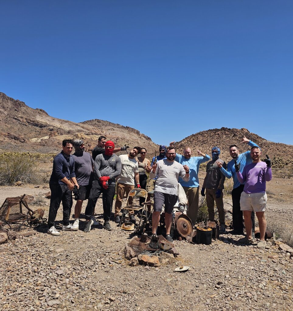 Twelve men pose in a Nevada desert ghost town near Las Vegas, with old mining machinery and mountains under a blue sky.