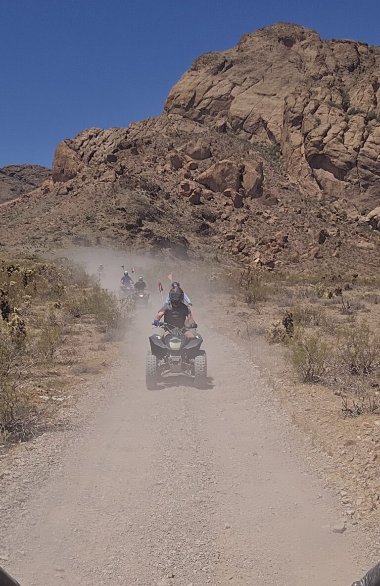 Group on an ATV tour rides dusty desert trails near Las Vegas, Nevada, with rocky hills and clear blue skies in the background.